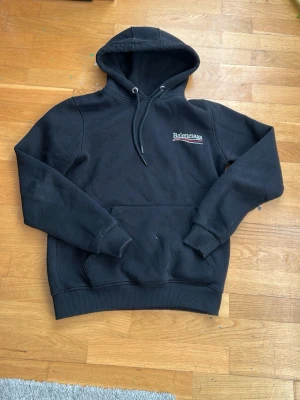 Svart Zara hoodie med tryck - Snygg svart hoodie från Zara med vit logga och röd-vit detalj på bröstet och ryggen. Klassisk känguruficka framtill, ribbade muddar och justerbar huva med snören. Perfekt för dig som gillar streetwear och vill sticka ut med ett exklusivt plagg. Ett liten hål i armhålan 