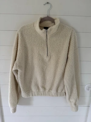 Beige teddytröja med halv dragkedja - Mysig beige teddytröja med halv dragkedja och hög krage. Tröjan har långa ärmar, resår vid midjan och är tillverkad i fluffigt, mjukt material som håller dig varm. Perfekt för kyliga dagar och enkel att matcha med jeans eller leggings.