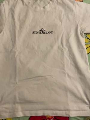 Vit Stone Island t-shirt - Vit t-shirt från Stone Island med klassisk logga tryckt på bröstet och stort kompassmärke på ryggen. T-shirten har rund hals och korta ärmar, tillverkad i mjuk bomull för en skön och avslappnad stil.