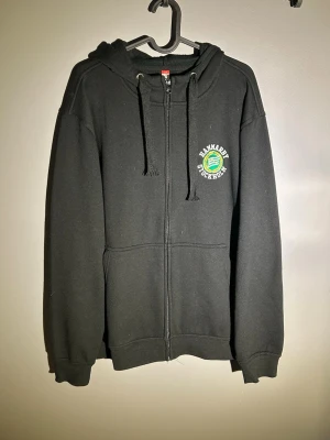 Hammarby hoodie - Svart hoodie med dragkedja och huva, samt snörning i halsen. Tryck med grön och vit logga på bröstet där det står HAMMARBY SOCKHOLM. Mjuk bomullskänsla och ribbade muddar vid ärmslut och nederkant.