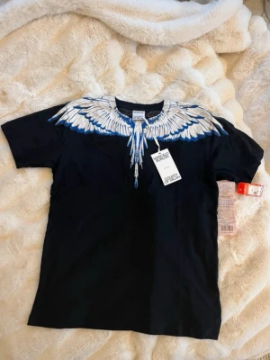 Svart t-shirt med blått vingmönster - Snygg svart t-shirt från Marcelo Burlon med ikoniskt vingmönster i vitt och blått runt halsen. T-shirten har korta ärmar och är gjord i mjuk bomull. Perfekt för dig som vill sticka ut med en unik och trendig look.