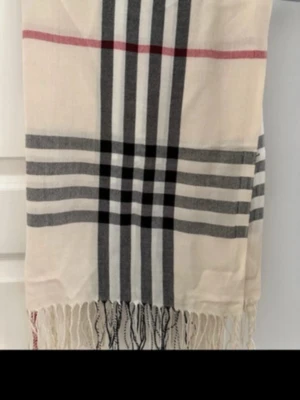 Beige halsduk med rutigt mönster - Stilren beige halsduk med klassiskt rutigt mönster i svart, vitt och rött. Halsduken har fransar i ändarna och är tillverkad i ett mjukt tyg som känns skönt mot huden. Perfekt accessoar för att lyfta din outfit.