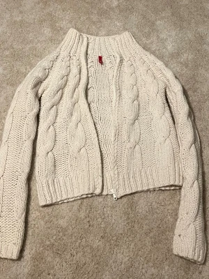 Beige kabelstickad kofta Divided - Säljer en beige kabelstickad kofta från Divided med dragkedja framtill. Koftan har grovstickat mönster, ribbade muddar och hög krage. Perfekt för dig som gillar en mysig och trendig look till hösten eller vintern.