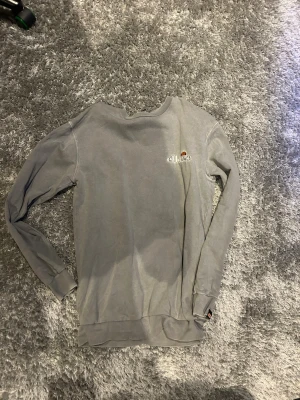 Grå sweatshirt från Ellesse - Säljer en grå sweatshirt från Ellesse med liten broderad logga på bröstet. Tröjan har rund halsringning, långa ärmar och ribbade muddar vid ärmslut och nederkant. Perfekt för en avslappnad och sportig stil.