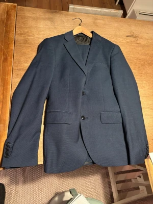 Mörkblå kostym från Lindbergh - Stilren mörkblå kostym från Lindbergh med två knappar framtill, klassiska slag och fickor med lock. Slim fit med storlek eu 46, jag hade S när jag använde den. Har använts 3-4 ggr så den är i mycket bra skick. Perfekt till student eller skolavslutning. Tveka inte att fråga efter fler bilder. 