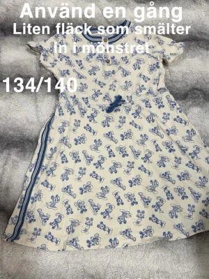 Vit och blå kortklänning med mönster - Söt kortklänning i vitt med blått blommigt mönster och blå knytband i midjan. Klänningen har korta ärmar, rund halsringning och dekorativa knappar framtill. Perfekt för varma dagar och har en avslappnad passform.🌸 Använd en gång. Finns en liten fläck som smälter in i mönstret. Storlek 134/140.