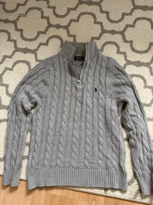 Grå kabelstickad tröja Polo Ralph Lauren - Kabelstickad grå tröja från Polo Ralph Lauren med halv dragkedja och hög krage. Tröjan har ribbade muddar och nederkant samt den klassiska blå logotypen broderad på bröstet. Perfekt för lager på lager och riktigt snygg till jeans.