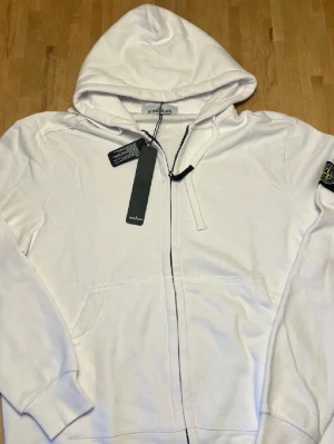 Vit hoodie från Stone Island - Snygg vit hoodie från Stone Island med dragkedja framtill och klassisk logopatch på ärmen. Hoodien har långa ärmar, fickor fram och justerbar huva med snören. Perfekt för en clean och stilren look.