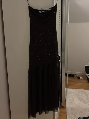 Brun långklänning med glitter GINATRICOT - Elegant svart långklänning med tunna axelband och glittriga detaljer över hela klänningen. Klänningen har en lös, svepande kjol och ett mjukt fall. Perfekt för dig som vill sticka ut med en stilren look. Klänningen är helt ny och har aldrig använts och pris lappen är kvar.
