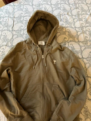 Olivgrön zip hoodie från Ami - Snygg olivgrön hoodie från Ami Alexandre Mattiussi med dragkedja framtill, två fickor och justerbar huva. Liten vit Ami-logga på bröstet. Tillverkad i mjuk bomull för skön känsla och avslappnad stil.