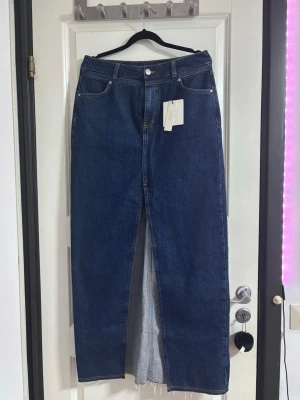 Mörkblå jeanskjol med slits - Snygg mörkblå lång jeanskjol med hög midja och slits framtill. Klassisk femficksmodell med rå kant nedtill och knapp- samt dragkedjestängning. Perfekt för dig som gillar en trendig och avslappnad stil.