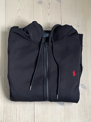 Svart hoodie från Polo Ralph Lauren - Svart hoodie med dragkedja från Polo Ralph Lauren. Klassisk design med huva och snörning samt röd broderad logga på bröstet. Perfekt för en avslappnad och stilren look. Använd i ca 2 månader men fortfarande bra skick storlek M