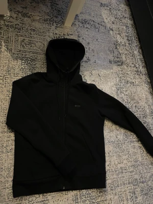 Hugo boss kofta  - Svart hoodie från BOSS med stilren logga på bröstet, hel dragkedja och snörning i huvan. Tröjan har två fickor framtill och är gjord i mjuk bomullsmix. Perfekt för chill dagar och streetwear vibes.