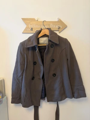 Brun kort rock från Zara - Kort brun rock från Zara med bred krage, dubbelknäppning och avtagbart midjebälte. Tillverkad i ett mjukt tyg som ger en snygg touch. Rocken har långa ärmar och stora svarta knappar framtill. Storleken kan dock kännas för liten.