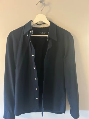 Mörkblå skjorta från Boulélid - Stilren mörkblå skjorta från Boundelid med klassisk krage och vita knappar. Skjortan har lång ärm och en skräddarsydd passform som ger en clean och modern look. Perfekt för dig som gillar minimalistisk stil.