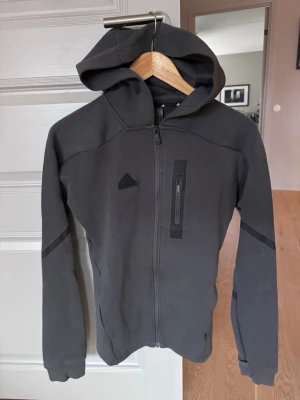 Mörkgrå hoodie från Adidas - Snygg mörkgrå hoodie från Adidas med dragkedja framtill och en bröstficka med dragkedja. Tröjan har huva och diskreta svarta detaljer på ärmarna. Perfekt för en sportig och avslappnad stil. Passar: vuxen herr XS vuxen dam S Junior M