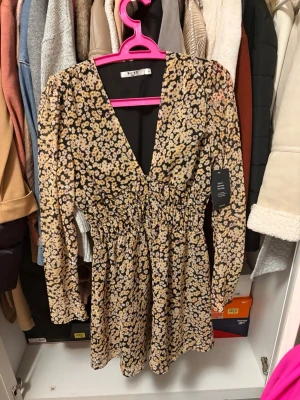 Blommig kortklänning från NA-KD - Supersöt kortklänning från NA-KD med blommigt mönster i beige, svart och gul. Djup v-ringning, långa ärmar och smockad midja som ger en snygg siluett. Perfekt för vår och sommar, lätt och luftig i polyester.