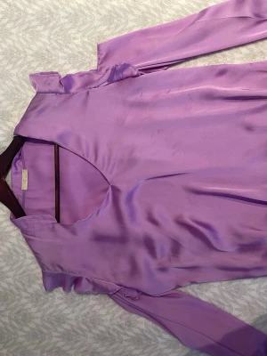 Lila sidenblus från E7 Paris - Snygg lila blus i glansigt sidenmaterial. Blusen har långa ärmar och en v-ringad halsringning med volangdetaljer vid axlarna. Storlek står ej men skulle säga s/m