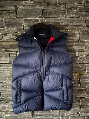Mörkblå dunväst från Tommy Hilfiger - Snygg mörkblå dunväst från Tommy Hilfiger med dragkedja framtill och två fickor på sidorna. Västen har en hög krage och diskret logga på bröstet. Perfekt för lager på lager och ger en sportig vibe. Materialet är dunfyllning för extra värme.