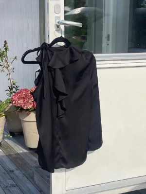Svart oneshoulder blus med volang - Snygg svart oneshoulder blus med volangdetalj längs halsringningen och knytning vid axeln. Blusen har en elegant och modern look, perfekt för att sticka ut. Tillverkad i ett mjukt, slätt tyg som faller fint.