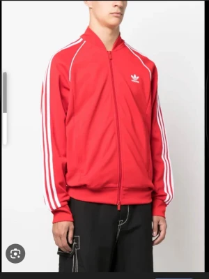 Röd Adidas vindjacka med vita ränder - Sportig röd vindjacka från Adidas med klassiska vita ränder längs ärmarna och vit Adidas-logga på bröstet. Jackan har dragkedja framtill, ribbade muddar och lång ärm. Perfekt för en streetwear-look.