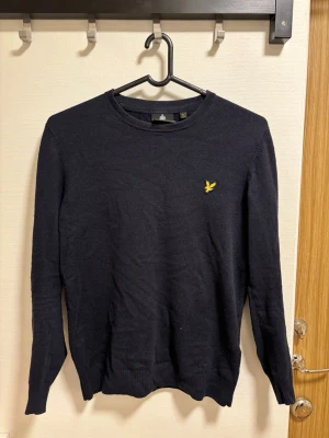 Mörkblå stickad tröja från Lyle & Scott - Snygg mörkblå stickad tröja från Lyle & Scott med klassisk rund hals och gul logotyp på bröstet. Tröjan har långa ärmar och ribbade muddar vid ärmslut och nederkant. Perfekt för dig som gillar stilrena plagg.