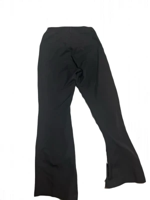 Svarta utsvängda kostymbyxor - Säljer ett par svarta kostymbyxor med utsvängda ben och hög midja. Byxorna har en klassisk look med diskret knappstängning och är sydda i ett mjukt, följsamt tyg som ger en snygg siluett. Perfekta för dig som gillar stilrena och trendiga plagg. Passar M-L