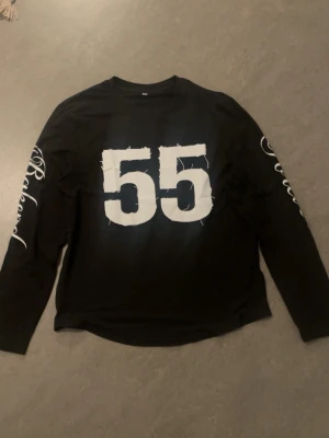 Svart långärmad t-shirt med tryck - Cool svart långärmad t-shirt med stort vitt tryck av siffran 55 på bröstet och text på ärmarna. Perfekt för dig som gillar sportiga och street-inspirerade plagg. Rund halsringning och relaxed fit.