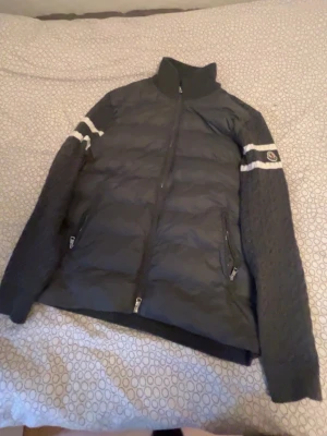 Mörkgrå Moncler cardiganjacka - Snygg mörkgrå cardiganjacka från Moncler med quiltad front och stickade ärmar. Jackan har vita ränder på ärmarna och Moncler-logga på vänster ärm. Dragkedja framtill och två fickor med dragkedja. Perfekt för lager-på-lager och streetstyle.