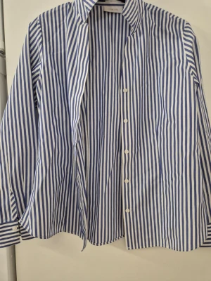 Skjorta - Klassisk skjorta från Chico's i blåvitt randigt mönster. Skjortan har lång ärm, knappar framtill och krage. Tillverkad i bomull för en skön och luftig känsla. Perfekt till jeans eller kostymbyxor för en clean look.