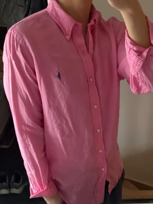 Rosa linneskjorta från Ralph Lauren - Klassisk rosa linneskjorta från Ralph Lauren med button-down krage och blå broderad logga på bröstet. Skjortan har lång ärm, normal passform och är tillverkad i 100% linne. Perfekt för en clean och stilren look. Där är några fläckar på armen som man ser på sista bilden men syns knappt om man inte kollar jättenoga och lite missfärgad på insidan av kragen men syns inte heller när man har den på