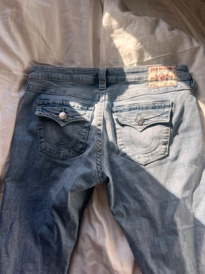 True Religion ljusblå bootcut jeans - Säljer ett par ljusblå jeans från True Religion med bootcut-modell. Jeansen har snygga bakfickor med lock och dekorativa sömmar samt silverfärgade knappar. Klassisk patch bak i midjan och mjukt denimtyg. Supersnygga och sparsamt använda! Säljes endast då de inte längre passar.