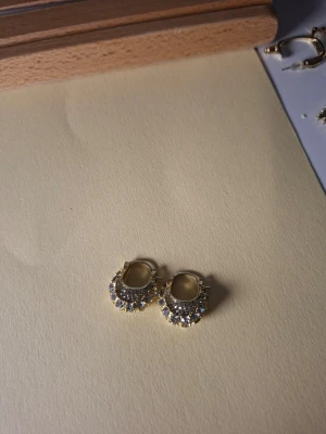 Guldpläterade creoler med vita stenar - Snygga små creoler i guldplätering med gnistrande vita stenar runt hela örhänget. De har en rund form och klicklås som gör dem enkla att ta på och av. Perfekta för dig som gillar lite extra bling i vardagen.