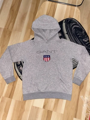 Grå hoodie från GANT med logga - Snygg grå hoodie från GANT med stor huva och klassisk GANT-logga broderad på bröstet samt ett sköldmärke i rött, vitt och blått. Kom med prisförslag!