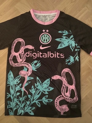 Inter Nike matchtröja svart/rosa - Unik Inter-matchtröja från Nike i svart med rosa och turkosa detaljer. Tröjan har tryckta ormar och blad, rosa ribbad krage och ärmslut samt klubbmärke och sponsorlogga framtill. Perfekt för dig som vill sticka ut på planen eller läktaren. perfekt till sommaren som streetwear