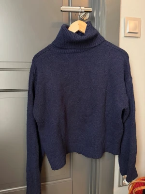 Mörkblå stickad polotröja - En mörkblå stickad polotröja med hög krage och långa ärmar. Tröjan har en avslappnad passform och ribbstickade detaljer vid kragen, ärmslut och nederkant. Perfekt för att hålla sig varm under kyliga dagar.