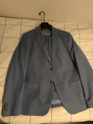 Ljusblå kavaj med två knappar - Snygg ljusblå kavaj med två knappar framtill och klassisk passform. Kavajen har två fickor med lock, bröstficka och dekorativa knappar vid ärmsluten. Perfekt för en stilren look. Materialet ser ut att vara en tunnare vävd textil.