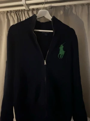 Mörkblå zip-tröja från Ralph Lauren - Mörkblå zip-tröja från Ralph Lauren med grönt broderat logotyp på bröstet, Storlek L men känns som M(14-16), priset kan diskuteras