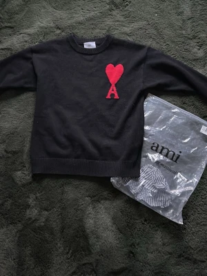 Svart stickad tröja från Ami - Svart stickad tröja från Ami Alexandre Mattiussi med röd hjärt- och A-detalj på bröstet. Tröjan har rund halsringning och ribbade muddar vid ärmslut och nederkant. Perfekt för dig som gillar stilrena plagg med en twist.