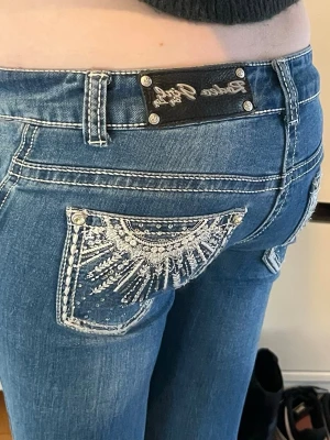 Blåa bootcut jeans från Rock & Republic - Säljer ett par blåa bootcut jeans från Rock & Republic med vita kontrastsömmar och snygg broderad bakficka med blingdetaljer. Jeansen har låg midja och klassisk femficksmodell. Perfekta för dig som gillar statement-jeans med unik design.