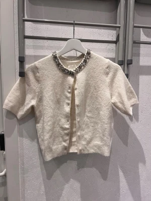 Vit kortärmad kofta med pärldetaljer - Supersöt vit kofta med korta ärmar och rund hals. Knäppning framtill och dekorerad med glittrande pärlor runt halsringningen. Mjuk, stickad känsla och ribbade kanter nedtill och vid ärmslut. Perfekt för att lyfta en enkel outfit.