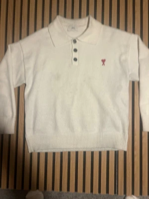 Vit stickad tröja från Ami Paris - Vit stickad tröja från Ami Paris med krage och tre knappar framtill. Tröjan har ett rött hjärtemblem med bokstaven A broderat på bröstet. Ribbstickade muddar vid ärmslut och nederkant ger en snygg finish. Passar M och L i storlek på herrar. Väldigt bra skick. Ifall ni har frågor skriv bara!!!