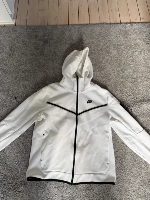 Vit Nike hoodie med svarta detaljer - Snygg vit hoodie från Nike med dragkedja och svarta detaljer längs dragkedjan och bröstet. Klassisk huva och två fickor framtill. Tillverkad i mjukt material som känns skönt mot huden. Nike-logga broderad på bröstet för en sportig look.