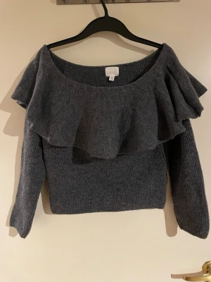 Mörkgrå stickad tröja med volang - Snygg mörkgrå stickad tröja med bred volang runt halsen och axlarna. Tröjan har lång ärm och en lite kortare, boxig passform. Perfekt för dig som gillar att sticka ut med detaljer. Materialet är mjukt och känns skönt mot huden.