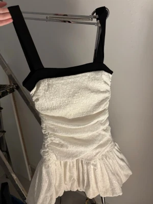 Vit kort klänning från Zara - Supersöt vit kort klänning från Zara med breda svarta axelband och volang nedtill. Klänningen har en rynkad struktur och markerad midja som ger en snygg siluett. Perfekt för sommarens alla tillfällen! Till och med prislappen kvar