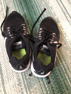 Nike Zoom Pegasus 34 svarta sneakers - Snygga svarta Nike Zoom Pegasus 34 sneakers med vit sula och mesh-överdel för bra ventilation. Skorna har svarta snören, vita detaljer och en grön innersula. Perfekta för dig som gillar sportig stil och komfort.