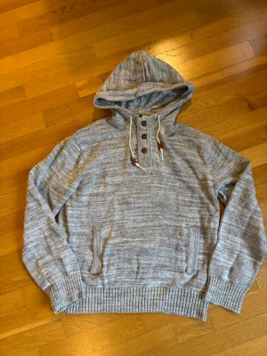 Ljusgrå stickad hoodie med knappar - Säljer en ljusgrå stickad hoodie med ribbade muddar, fickor framtill och knappar vid halsen. Tröjan har en stor huva med dragsko och är perfekt för dig som gillar en avslappnad och stilren look. Materialet är mjukt och skönt.