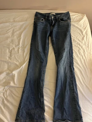 Blå bootcut jeans med fickdetaljer - Säljer ett par klassiska blå bootcut jeans med låg midja och snygga fickdetaljer bak. Jeansen har en lätt tvättad look och är tillverkade i denim. Perfekta för dig som gillar en avslappnad men ändå trendig stil. Byxorna är från Gina tricot och har mycket små slitningar längst ner