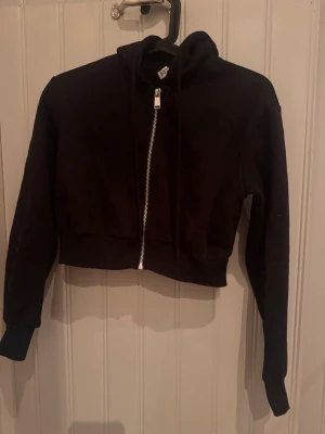 Svart cropped hoodie från H&M Divided - Säljer en svart cropped hoodie från H&M Divided. Hoodien har dragkedja framtill, huva med dragsko och långa ärmar. Perfekt till ett avslappnat streetwear-look. Tillverkad i mjukt bomullsmaterial för skön känsla.