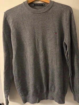 Grå stickad tröja Tommy Hilfiger - Klassisk grå stickad tröja från Tommy Hilfiger med rund halsringning och diskret logga broderad på bröstet. Tröjan har långa ärmar och ribbade muddar vid ärmslut och nederkant.  Stolek M/L